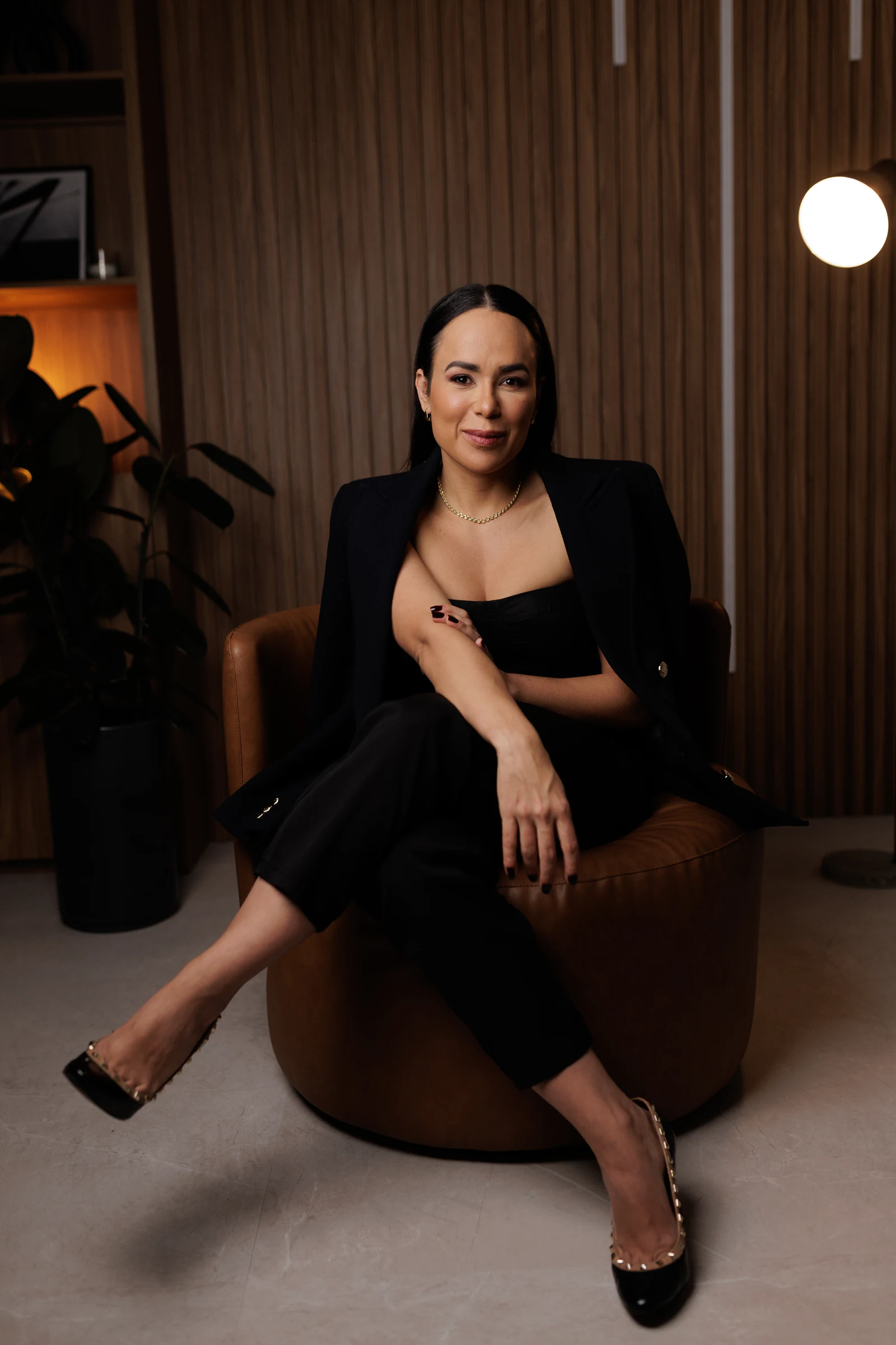 Dra. Paula Fernandes de blazer preto, sentada em poltrona de couro caramelo no consultório