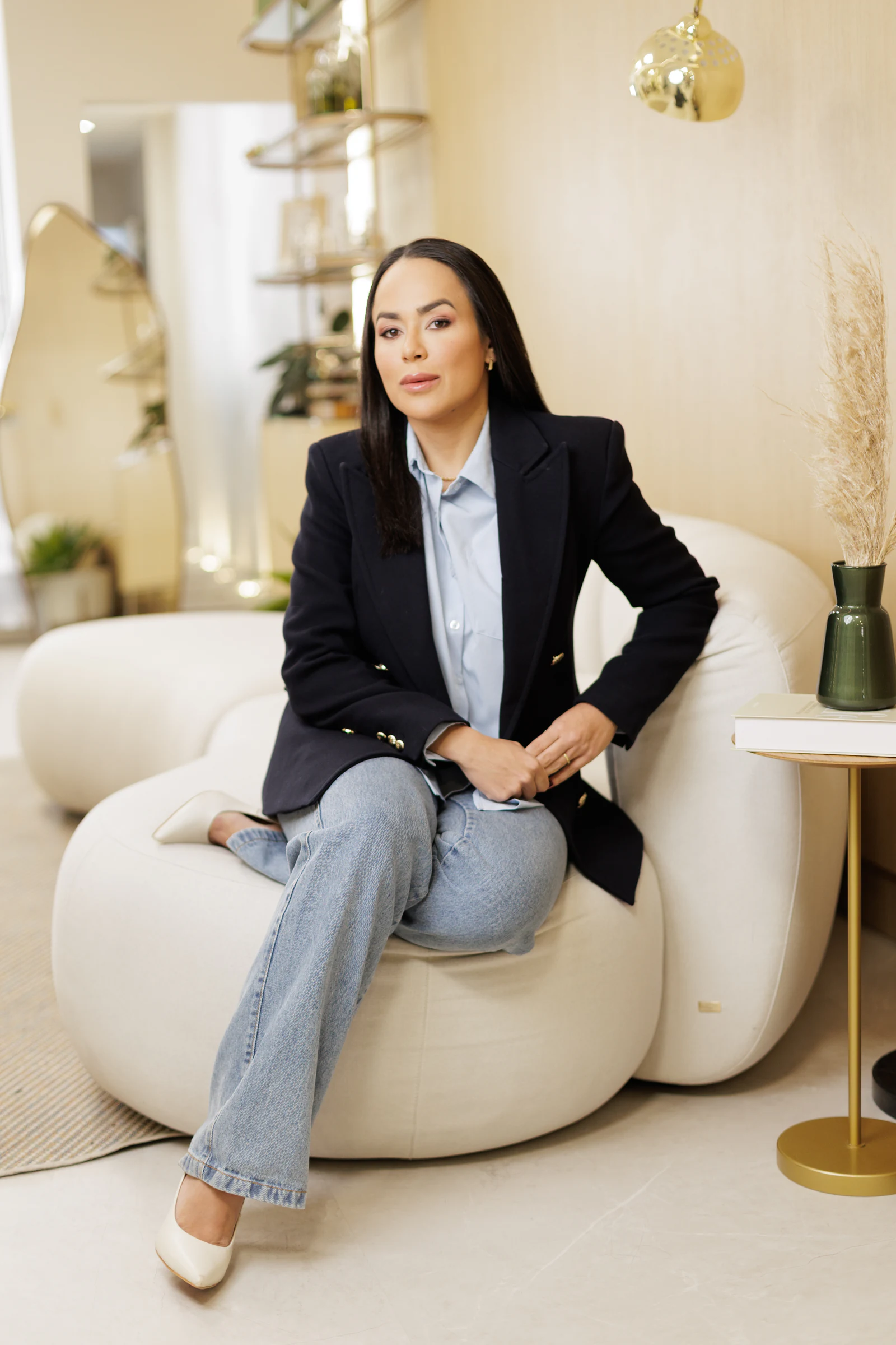 Dra. Paula Fernandes de blazer preto e jeans, sentada em um sofá claro no consultório